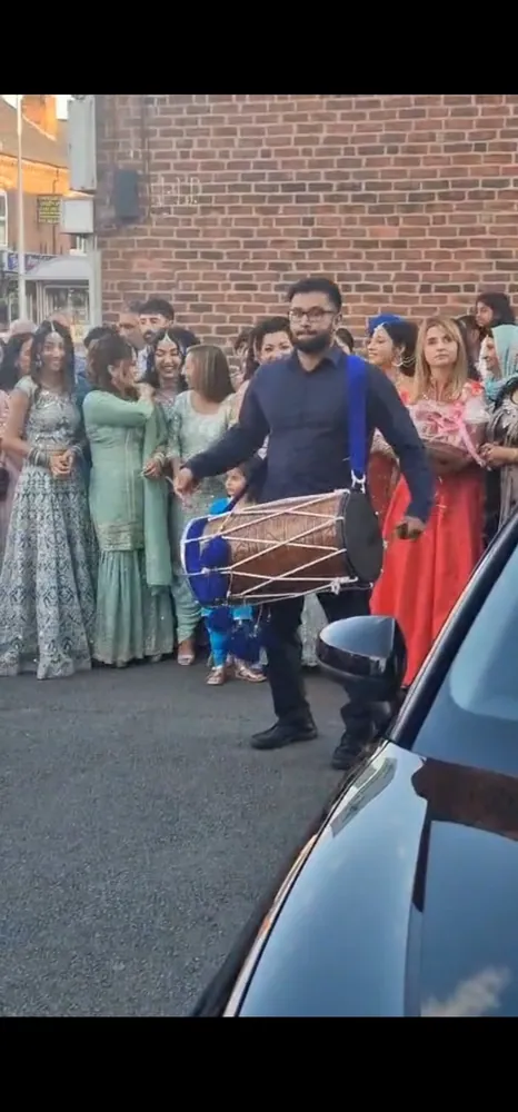 Manraj playing dhol at a baraat, wedding guests gathered behind him